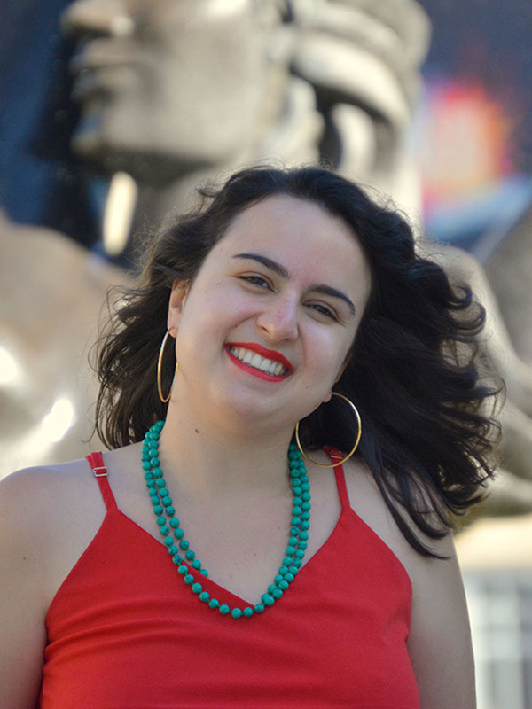 A headshot of Bruna Kalil Othero, who wears a red dress and poses in front of the Showalter Fountain.
