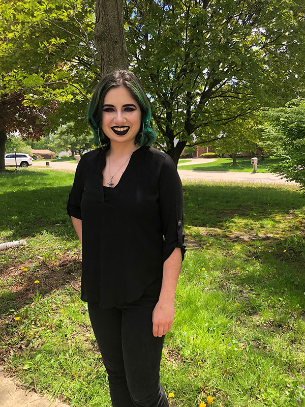 A headshot of Mariah Cardenas, who wears black pants and a black shirt and poses outside.