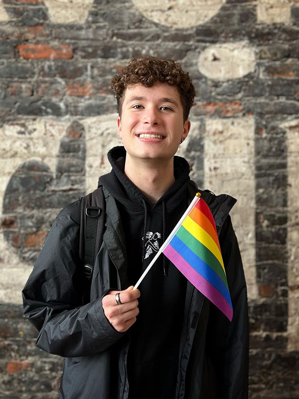 A headshot of Isaac Babb, who wears a dark hoodie and poses with a pride flag.