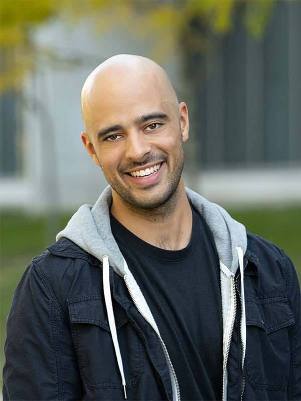 A headshot of Santiago Arroniz, who wears a dark blue jacket with a gray hoodie and poses outdoors.