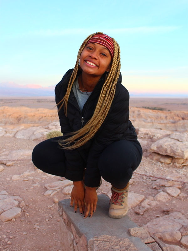 Trinity Barnes poses in a rocky dessert, with the sun setting in the background.