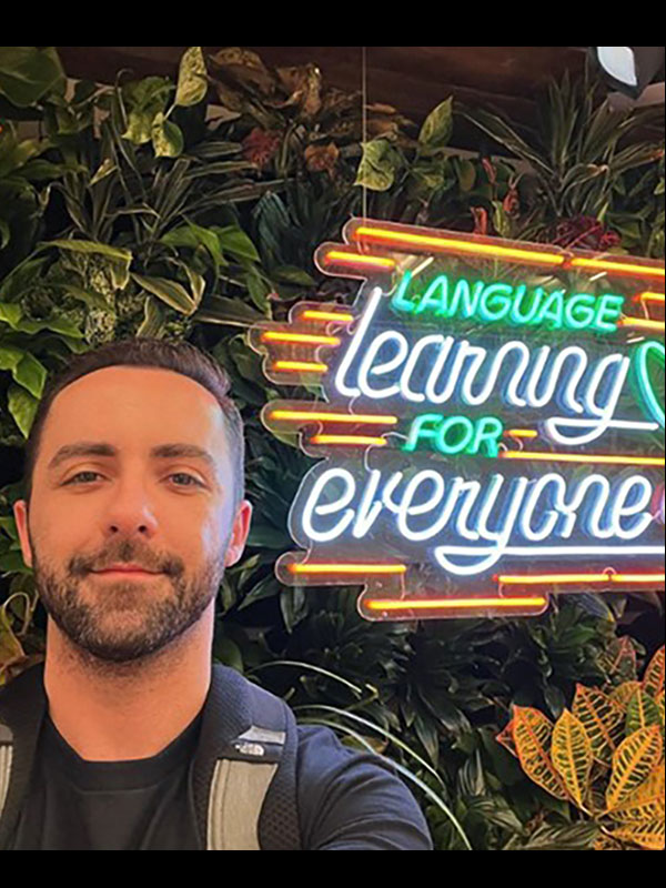 A headshot of Fernando Melero Garcia, who poses beside a sign that says, "Language learning for everyone."