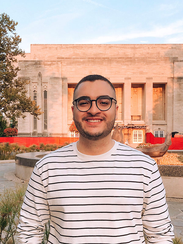 A headshot of Leandro Martan, who wears a white, striped shirt and poses in front of Showalter Fountain.