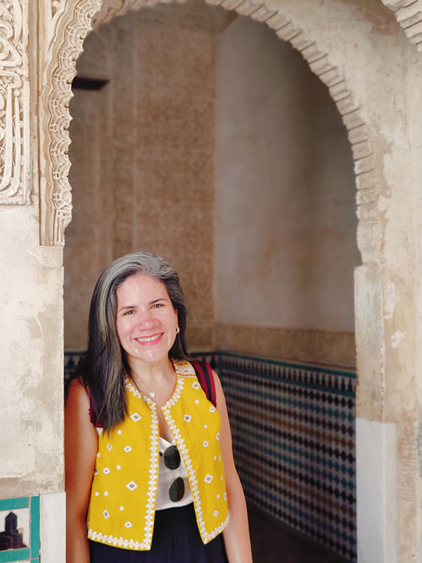 A headshot of Monica Vega, who wears a yellow vest and poses in an archway.