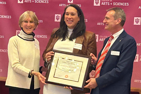 Three people standing in front of a College of Arts and Sciences backdrop as one receives a framed certificate.