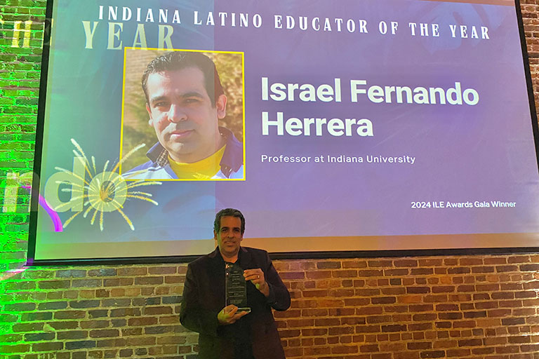 Person holding an award in front of a projected screen announcing them as the Indiana Latino Educator of the Year.