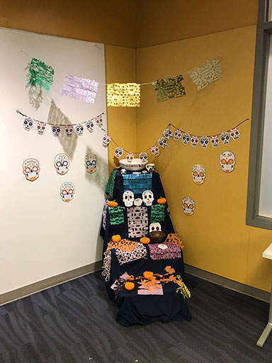 A decorated corner display with colorful paper cutouts, sugar skulls, and small pumpkins celebrating Día de los Muertos.