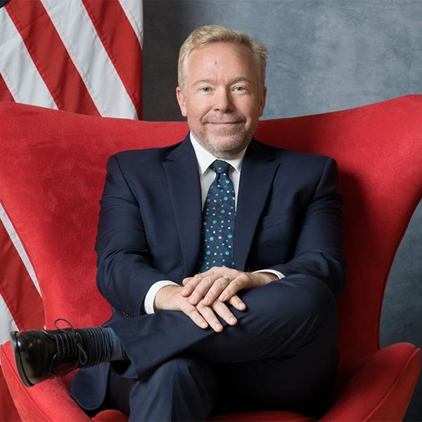 Person sitting cross-legged in a red chair wearing a navy suit and smiling, with a U.S. flag in the background.