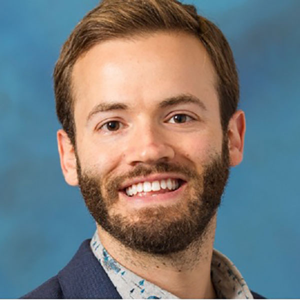 Portrait of a person with short hair and a beard smiling against a blue background.