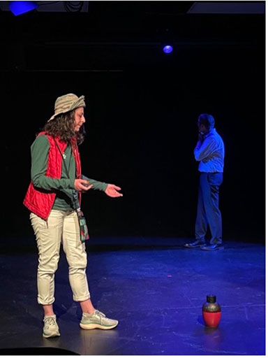 Two actors performing on stage under spotlight, one gesturing mid-scene while the other stands in the background.